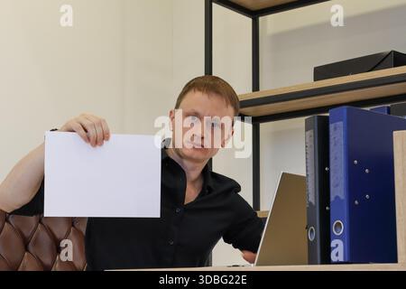 A young adult male in black shirt at office desk with laptop and folders, extending arm to hold blank white sheet sideways towards viewer. Stock Photo