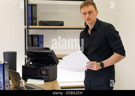 Adult caucasian man holds single blank paper near open printer in bright workspace with tech and storage. Stock Photo