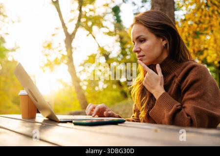 Woman freelancer working on laptop outdoors in autumn city park. Stock Photo