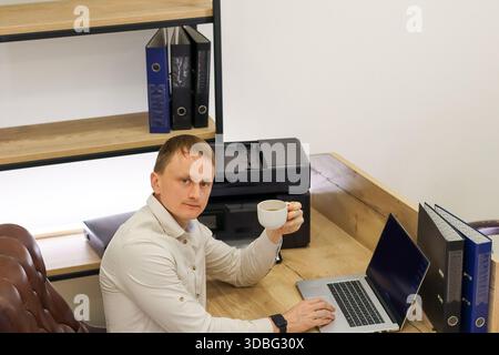 Office worker multitasks by working on portable computer and sipping from cup, near printer and file organizers in clean workspace. Stock Photo