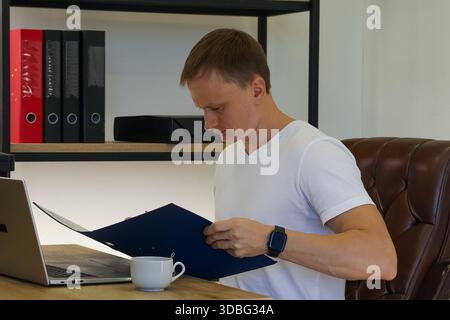 Close side view of a man in casual attire leaning forward to read from an open blue folder on his desk, with a smartwatch on wrist, laptop nearby, and Stock Photo