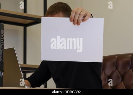Professional hiding face behind held white blank paper in workspace, seated at computer desk Stock Photo