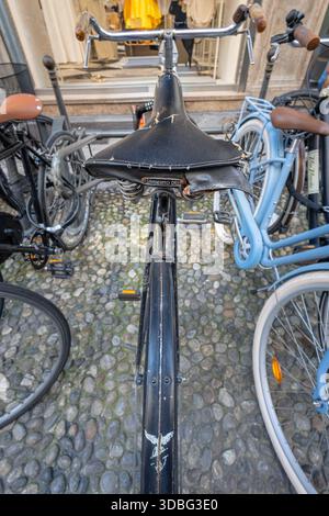 MILAN, ITALY, 06.19.2025:A close view of an aged bicycle saddle with peeling leather, surrounded by parked bikes on a cobblestone street. Urban scene Stock Photo