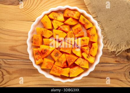 Pumpkin cut into pieces with sugar and cinnamon in a ceramic dish, ready to bake on a wooden table, close-up, top view. Stock Photo