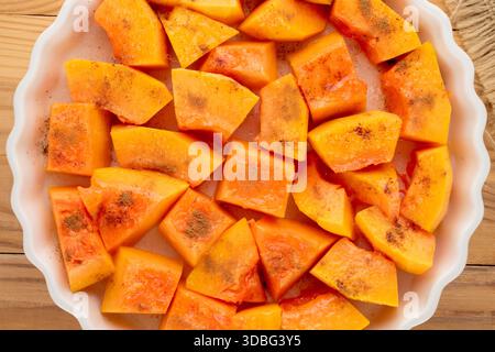 Pumpkin cut into pieces with sugar and cinnamon in a ceramic dish, ready to bake on a wooden table, close-up, top view. Stock Photo