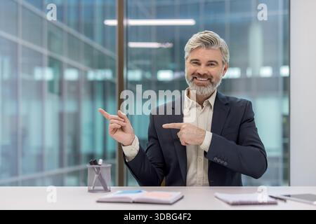 Experienced businessman sitting at an office desk, smiling confidently and gesturing to the left with both index fingers, indicating space for text or graphics Stock Photo