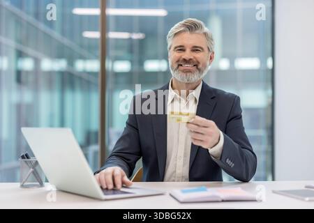 Mature businessman with a credit card smiling at the camera, using a laptop for online shopping and making secure e-commerce payments in a modern office setup Stock Photo