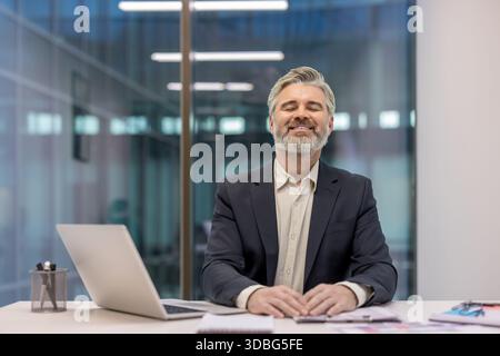 Mature businessman closing eyes, smiling, and relaxing at his office desk, enjoying a moment of peace and satisfaction while managing work stress and promoting mental wellness Stock Photo