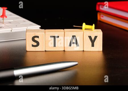Wooden blocks spell out STAY on a desk. A pen and computer are nearby, suggesting a work environment. Push pins add a touch of visual flair. Stock Photo
