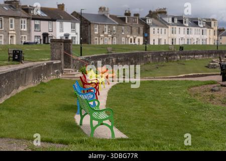 Vibrant rainbow benches along a grassy path by a stone seawall in a coastal town. Stock Photo