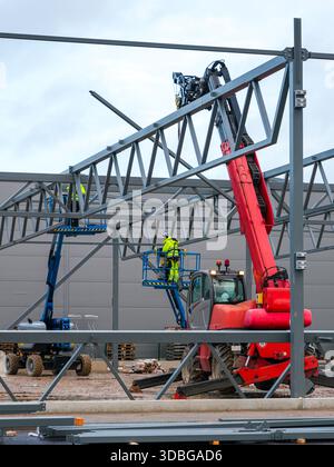Construction workers in safety gear assemble a steel framework using a crane and aerial lifts at an industrial building site Stock Photo