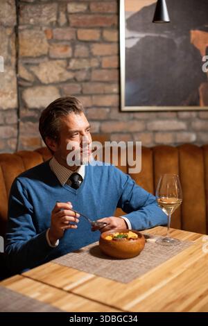 A man sits at a wooden table in a restaurant. He holds a spoon and looks thoughtful while eating. A glass of white wine sits nearby. The place has bri Stock Photo