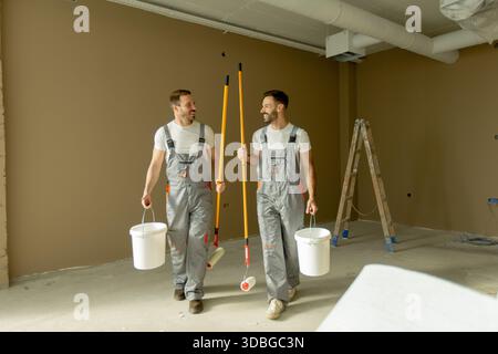 Two men walk together in an empty room carrying painting buckets and tools. They appear to be collaborating on a renovation task. The walls are painte Stock Photo