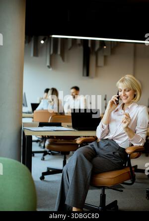 A woman is sitting in an office chair, talking on the phone. Several people are working at computers in the background. The office has a modern design Stock Photo