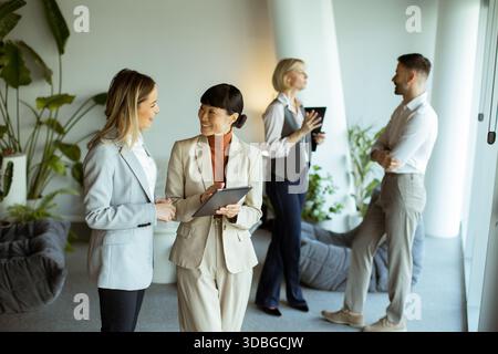 Two women talk and look at a tablet in a bright office. Two others stand nearby, engaging in conversation. Plants and soft seating are in the backgrou Stock Photo