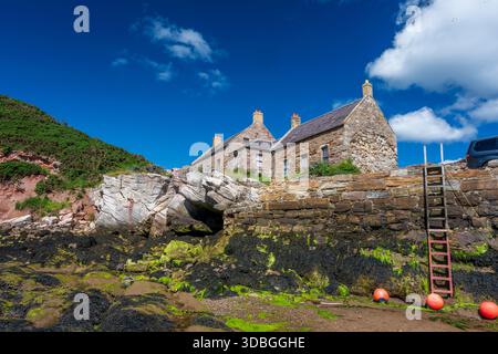 Stone cottages overlook a rocky coast with blue sky, calm sea, and green moss on the wall. Stock Photo