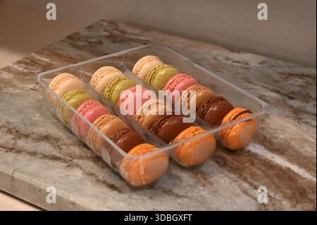 A collection of macarons in different colors and flavors. A clear plastic box filled with colorful assorted macarons on a textured stone. Stock Photo