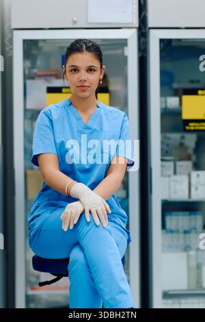 A medical professional is seated in a clinical environment, wearing blue doctor and gloves, ready to assist with patient care or medical tasks in a Stock Photo