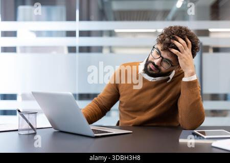 Bearded man sitting at his desk with a laptop, holding his head in a gesture of frustration and exhaustion, feeling overwhelmed by work stress and impending deadlines Stock Photo