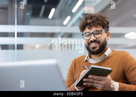 Smiling young man with curly hair and glasses taking notes in a bright modern office, holding a notebook and pen while looking at the camera, focused, professional and approachable Stock Photo