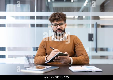 Young male employee in glasses attending a corporate meeting, actively listening and jotting down important information in a notebook at his desk in a contemporary office workspace Stock Photo