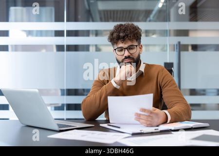 Young businessman analyzing financial reports and legal paperwork at a contemporary office desk, demonstrating focus and concentration during his work day Stock Photo