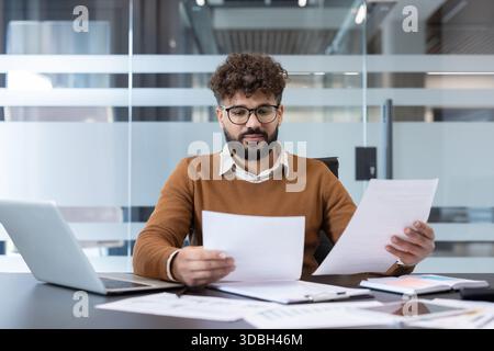 Young man analyzing important business documents and financial reports while concentrating at his desk in a modern corporate office, symbolizing hard work and analysis Stock Photo
