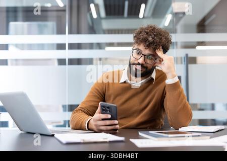 Young man in glasses sitting at office desk, appearing bored and distracted while using a mobile phone instead of working, feeling unmotivated and delaying tasks Stock Photo
