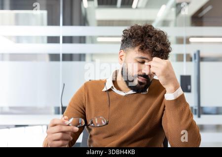 Young man experiencing burnout and stress at the office, feeling tired and having a headache while holding his eyeglasses, emphasizing workplace exhaustion and discomfort Stock Photo