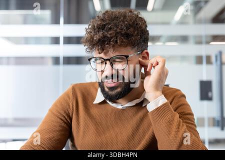 Man experiencing discomfort and pain, rubbing an aching ear while closing his eyes in an office setting, depicting health issues impacting concentration and well-being Stock Photo