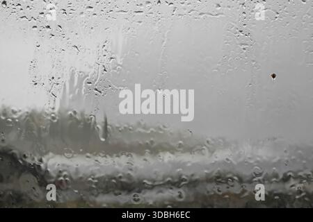 Close-up of a windowpane with heavy rain pouring down. Rainwater runs down the glass, leaving traces on the pane as the raindrops fall. Blurring the o Stock Photo