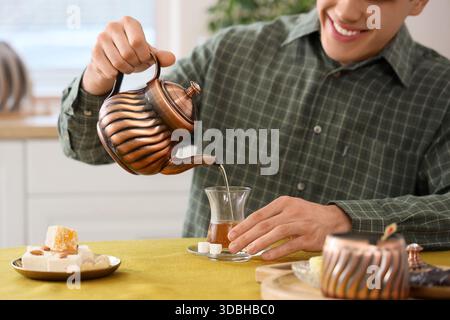Young man pouring Turkish tea into glass at table in kitchen Stock Photo