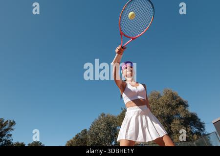 Woman tennis athlete hits ball with racket outdoors on sunny day, low angle view, blue sky copyspace, sport motion Stock Photo