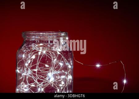 A glass jar filled with glowing string lights standing on a dark red surface, creating a cozy and festive holiday atmosphere. Stock Photo