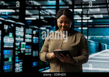 Smiling admin using tablet in data center to track metrics of gear used to host websites. Happy server room IT expert upgrading capacity of storage rackmounts running companies databases Stock Photo