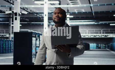 Technician walking in data center, configuring storage devices for minimal latency in AI workloads. African american employee using device in server farm to build neural networks, camera A Stock Photo