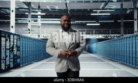 Cheerful admin walking in data center workplace, checking gear for system stability. Jolly IT professional overseeing server hub, configuring racks, optimizing performance, camera A Stock Photo