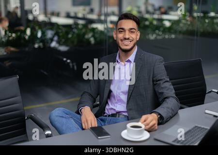 Young Man Sits in Office With Coffee and Smiles While Wearing a Suit and Casual Clothes Stock Photo