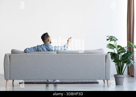 Man Sits on Couch Adjusting Air Conditioner in Bright Living Room While Relaxing on Sofa Stock Photo