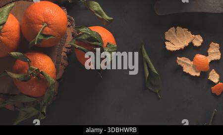 Overhead shot of fresh, wet tangerines with stems and leaves resting on a rustic wooden board, contrasted against a dark, textured slate or chalkboard Stock Photo