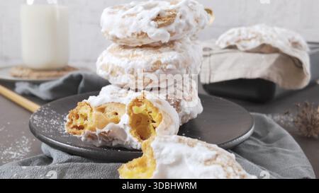 A vertical stack of fluffy, golden donuts covered in thick white sugar glaze sits on a dark plate. The airy interior of a broken donut is visible in t Stock Photo