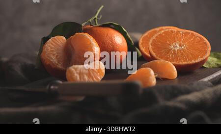 A beautifully dark and moody still life showcases fresh, whole tangerines with green leaves, alongside peeled segments and half-cut fruit, resting on Stock Photo