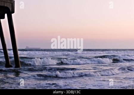 Ocean waves roll beneath a pier at dusk in Huntington Beach, California, with soft pastel skies and calm surf along the coast. Stock Photo
