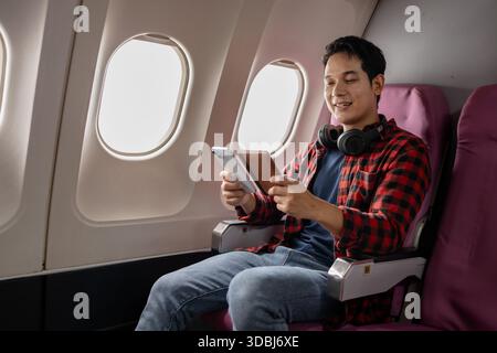 Asian man sitting comfortably on airplane seat near window, holding smartphone and passport, wearing headphones around neck, enjoying in-flight entert Stock Photo