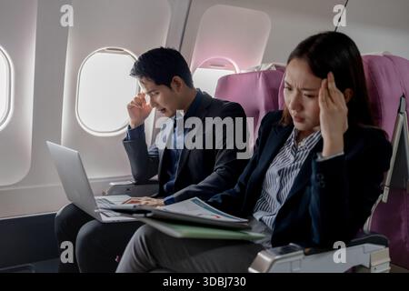 Two stressed Asian business professionals sitting on an airplane, holding their heads in frustration. The man looks at the laptop screen while the wom Stock Photo