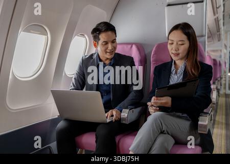 Two Asian business professionals are sitting next to each other on an airplane. The man is working on a laptop while the woman is holding a clipboard Stock Photo
