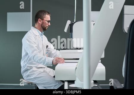 Caucasian middle aged man wearing lab coat operating advanced medical imaging device in clinical setting, focusing on screen Stock Photo