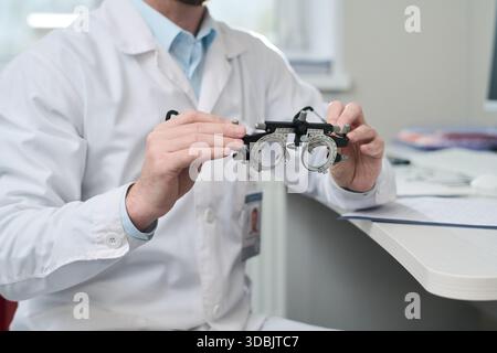 Middle aged Caucasian man wearing lab coat holding optometry trial frame with both hands, sitting at desk in medical office, preparing for vision test, face not visible Stock Photo