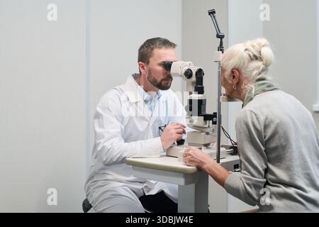 Caucasian middle aged male doctor performing eye examination on Caucasian senior woman using slit lamp biomicroscope in medical clinic, both subjects focusing on diagnostic procedure Stock Photo