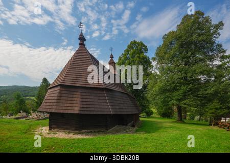 View of a rustic wooden church standing proudly on a grassy hill, its dark brown shingles contrasting against the bright blue sky, Topola, Presovsky k Stock Photo
